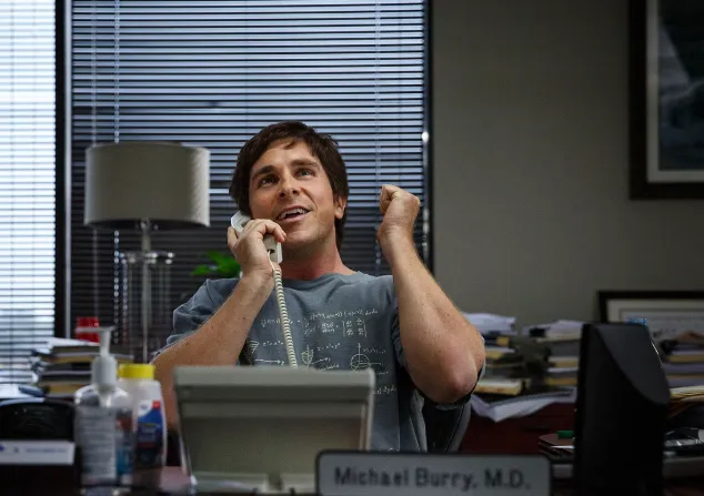 Christian Bale as Michael Burry in The Big Short, on the phone and pumping his fist behind a desk nameplate reading “Michael Burry, M.D.”